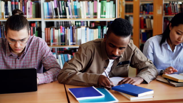 Schwenken-Sie-Schuss-Multi-ethnischen-Gruppe-konzentrierte-Studenten-Vorbereitung-für-die-Prüfung-beim-Sitzen-am-Tisch-in-der-Universitätsbibliothek