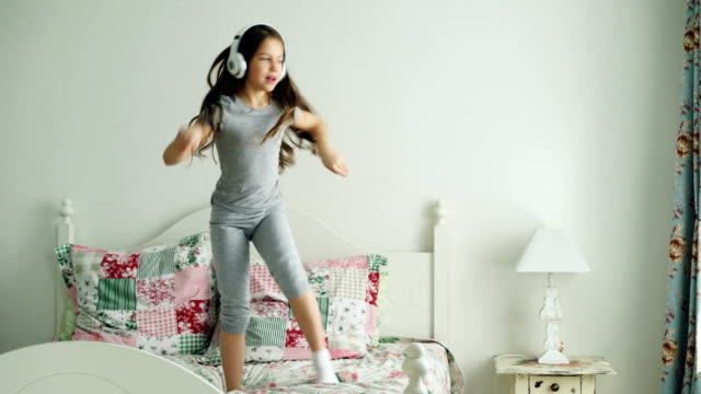 Funny-cute-little-girl-in-wireless-headphones-dancing-and-have-fun-on-bed-at-home-in-cozy-bedroom