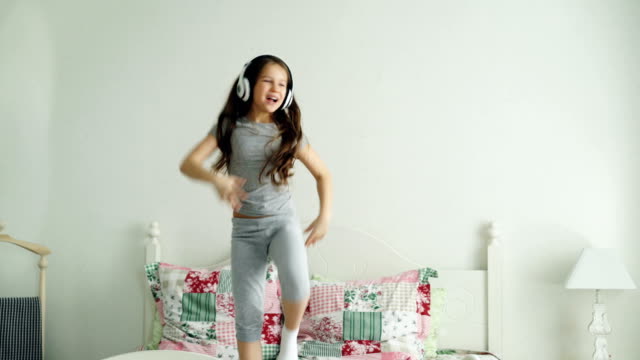 Funny-cute-little-girl-in-wireless-headphones-dancing-and-have-fun-in-holiday-morning-jumping-on-bed-at-home-in-cozy-light-bedroom