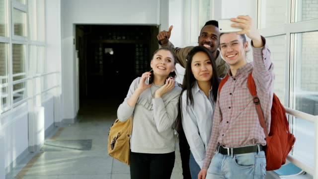 Gruppe-von-multi-ethnischen-Studenten-nehmen-Selfie-auf-Smartphone-Kamera-stehend-im-Flur-der-Universität.-Hipster-Mann-hält-Telefon-und-Freunde-stellen-positiv