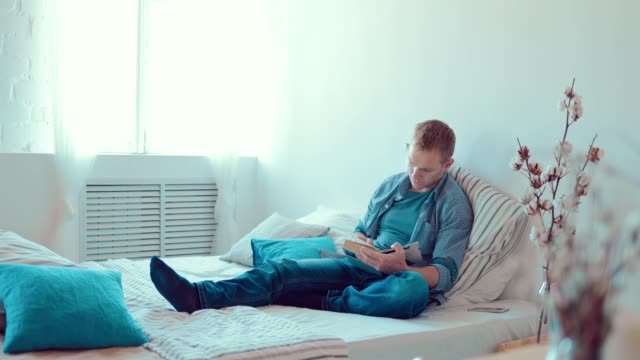 Young-man-reading-book-sitting-on-sofa-at-home