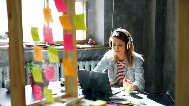 Pretty-blond-girl-is-listening-to-music-and-working-with-laptop-in-nice-loft-office.-She-is-making-notes-in-notepad,-smiling-and-dancing.-Relaxing-in-workplace-concept.