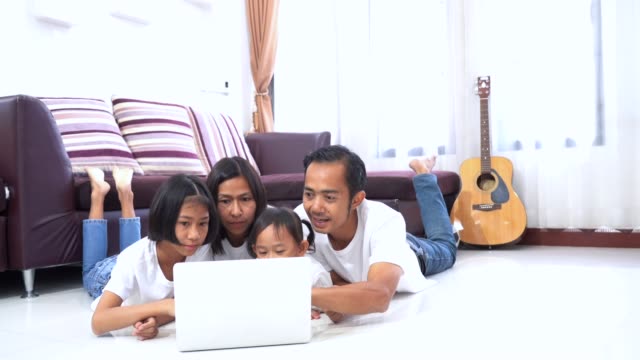 Feliz-familia-asiática-pequeña-hija-miente-en-una-planta-en-casa-y-utilizando-un-ordenador-portátil