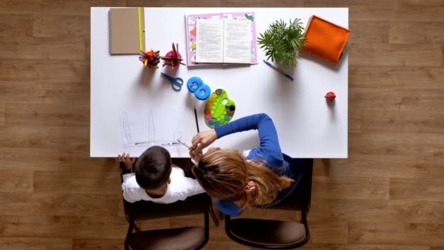 Junge-Mutter-mit-Sohn,-sitzen-hinter-Tisch,-Kind-macht-Hausaufgaben-Top-Shot-Parkett-Holzboden-malen