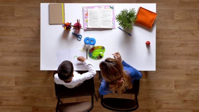 Junge-Mutter-mit-Sohn,-Kind,-erklären-hinter-Tisch-sitzen,-junge-tun-Hausaufgaben-Top-shot-Parkett-Holzboden