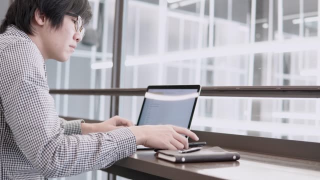 Junge-asiatische-Geschäftsmann-mit-Laptop-und-Smartphone-auf-hölzernen-Schreibtisch-im-Arbeitsraum.-Männliche-Hand-auf-Laptoptastatur-tippen.-Freiberuflicher-Lebensstil-im-digitalen-Zeitalter-Konzept.
