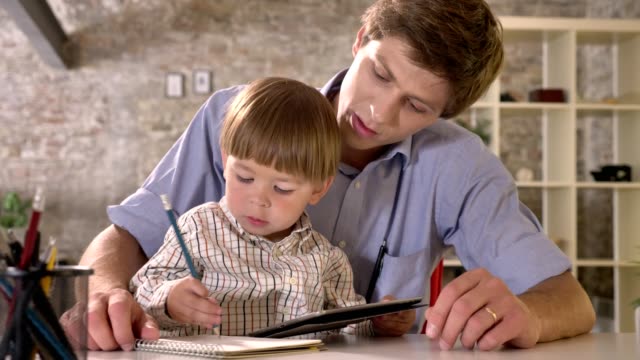 Little-boy-sitting-with-his-young-father-at-table-and-writing-on-paper,-looking-at-tablet,-modern-office-background