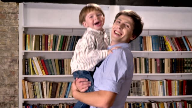 Junger-Vater-hält-seinen-kleinen-Sohn-und-springen-in-Bibliothek,-lächelnd-und-glücklich