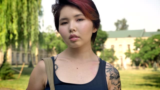 Portrait-of-young-asian-female-student-with-tattoo-standing-with-backpack-and-looking-in-camera-in-park-near-university,-confident