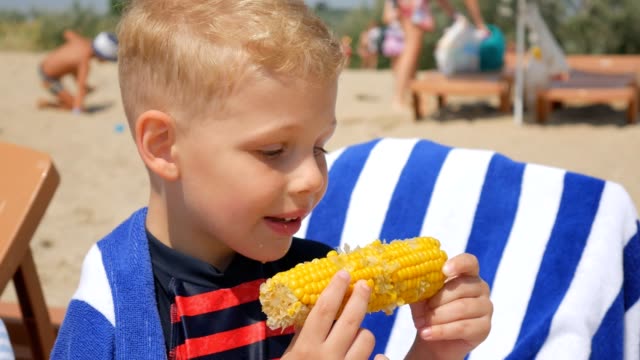 Little-hungry-boy-eating-corn-on-the-beach-of-the-sea.-Focus-on-the-boiled-corn.-Close-up.