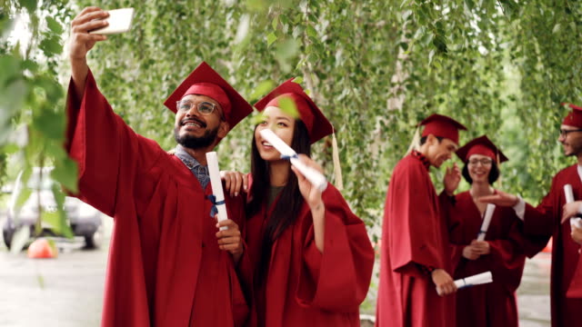 Two-mixed-race-students-are-taking-selfie-with-graduation-diplomas-wearing-mortarboards-and-gowns,-guy-is-holding-smartphone-and-taking-picture.