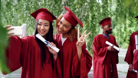 Two-attractive-girls-fellow-students-are-taking-selfie-on-graduation-day-holding-diplomas,-young-women-are-smiling,-posing,-making-hand-gestures-and-funny-faces.