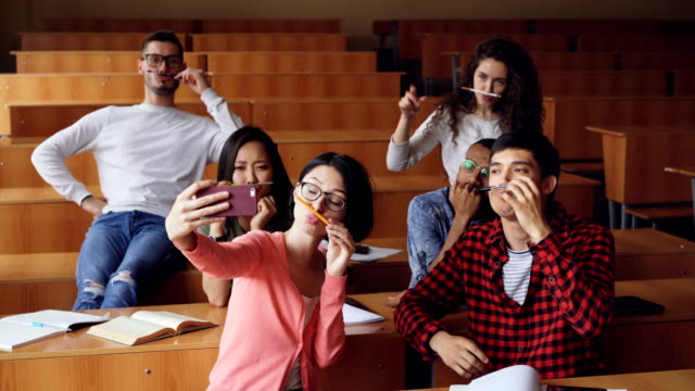 Verspielte-junge-Menschen-nehmen-lustige-Selfe-mit-Kugelschreiber-und-Bleistifte-als-Schnurrbart,-posieren-und-Handgesten-Daumen-nach-oben-zeigen-und-schlägt-im-Klassenzimmer-sitzen.