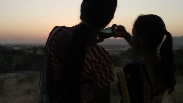 Two-women-silhouette-against-the-setting-sun-mobile-phone-photo-video-teamwork-romantic-viewpoint-love-bonding-together-tourist-rural-setting-friends-handheld-stabilized-lovers-follow--escape-surreal