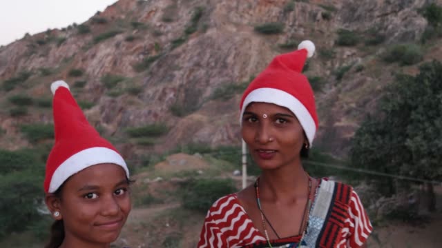 Zwei-Frauen-in-Santa-Hüte-und-traditionellen-indischen-Sari-helles-Muster-buntes-Kleid-Sommer-tropischen-auf-einem-Hügel-oben-Umarmung-Anteil-Punkt-Finger-schauen-Sie-direkt-auf-Kamera-Sonnenuntergang-Silhouette-lachen-Spaß-Freude-handheld
