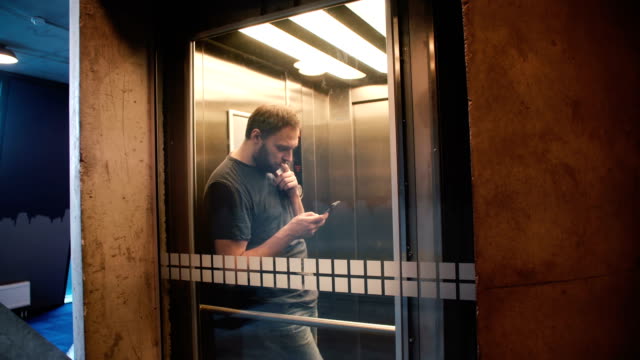 Young-happy-casual-man-riding-transparent-glass-elevator,-door-opens-and-he-walks-out-using-smartphone-shopping-app