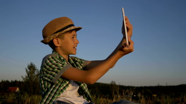 boy-in-a-hat-sits-on-top-and-talks-on-video-communication-using-a-tablet,-outdoors