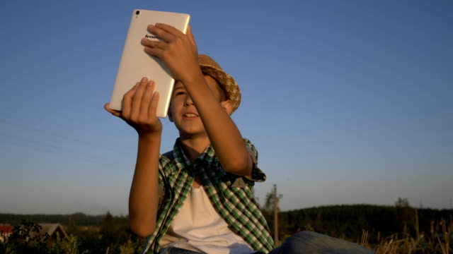 boy-in-a-hat-sits-on-top-and-talks-on-video-communication-using-a-tablet,-outdoors