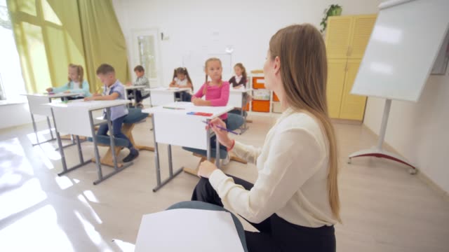 portrait-of-smiling-teacher-female-woman-during-education-lesson-with-pupils-in-classroom-at-Primary-school-on-unfocused-background