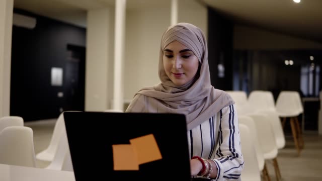 Smiling-confident-muslim-female-wearing-a-hijab-sitting-on-a-white-chair-in-the-modern-conference-hall-online-with-a-laptop-on-the-white-desk
