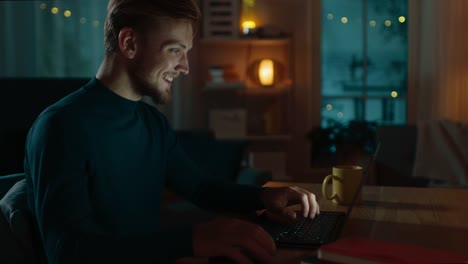 Abend-zu-Hause:-Portrait-of-Handsome-Man-sitzt-an-seinem-Schreibtisch-auf-einem-Laptop-arbeiten.-Lächelnde-Freelancer-arbeiten-am-Computer.-Richtungskontrolle-Ansicht.