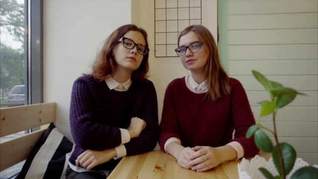 Two-pretty-girls-students-glasses-sitting-at-the-table-in-cafe-looking-at-camera