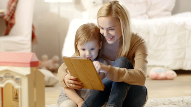Hermosa-madre-y-su-pequeña-hija-tienen-buen-tiempo-leyendo-libros-infantiles-en-una-tableta-de-la-computadora.-La-habitación-de-los-niños-está-llena-de-juguetes.
