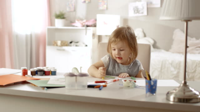 Linda-niña-se-sienta-en-su-mesa-y-dibuja-con-crayones.-Su-habitación-es-rosa,-dibujos-bonitos-colgando-de-las-paredes,-muchos-juguetes-por-ahí.
