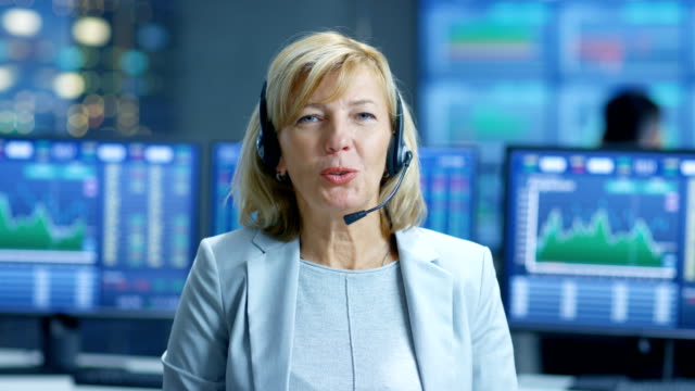 Chief-Sales-Force-Representative-Talks-into-the-Headset.-Behind-Her-People-Working,-Screens-Show-Stock-Market-Ticker-Numbers-and-Graphs.