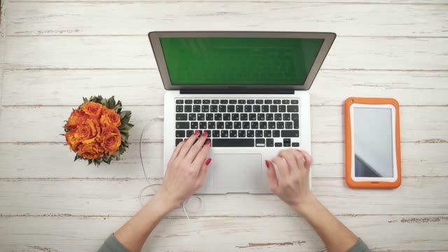Frau-freier-fügt-ein-Kopfhörer-in-einen-Laptop,-stehend-auf-dem-weißen-Holztisch-mit-Blumen-und-orange-Tablette,-und-arbeitet-darauf.-Ansicht-von-oben.-Hände-hautnah