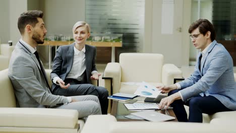 Panning-shot-of-businessman-discussing-financial-reports-with-female-and-male-business-partners-sitting-on-sofa-in-modern-office-hall-with