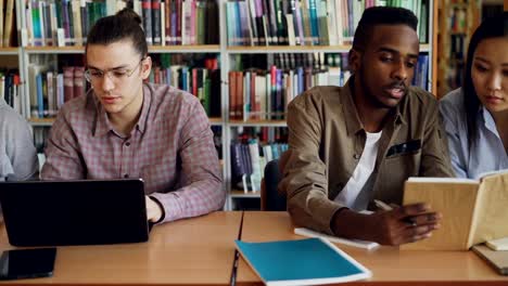 Pan-Schuss-von-Multi-ethnischen-Gruppe-konzentrierte-sich-Studenten-sprechen-und-Vorbereitung-für-die-Prüfung-beim-Sitzen-am-Tisch-in-der-Universitätsbibliothek