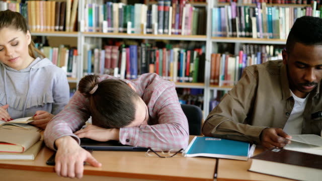 Pan-Schuss-von-fleißigen-Studenten-Prüfungsvorbereitung-tun-Hausaufgaben-und-müde-Kerl-schlafen-auf-Tabelle-in-der-Universitätsbibliothek