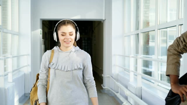 Beautiful-attractive-female-student-wearing-headphones-is-going-down-corridor-meeting-her-groupmates-giving-five-to-her-friend-and-moving-on.-She-show-middle-finger-to-african-american-guy