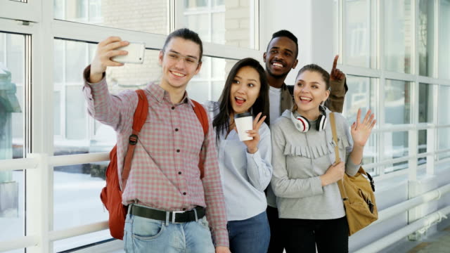 Grupo-de-cuatro-estudiantes-hombres-y-mujeres-positivos-multiétnicos-está-de-pie-en-el-amplio-corredor.-Tripa-de-hipster-tiene-smartphone-haciendo-selfie-de-todos