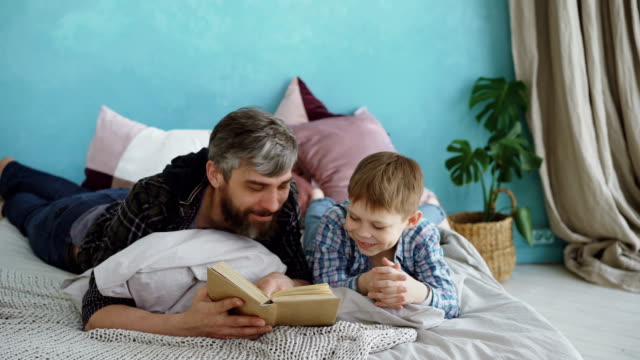 Fürsorglicher-Vater-ist-lustig-Buch-zu-seinem-Kind-zu-lesen,-während-junge-lacht-und-im-Gespräch-mit-seiner-Eltern.-Full-Size-Bett,-hellen-Kissen-und-Grünpflanzen-sind-sichtbar.