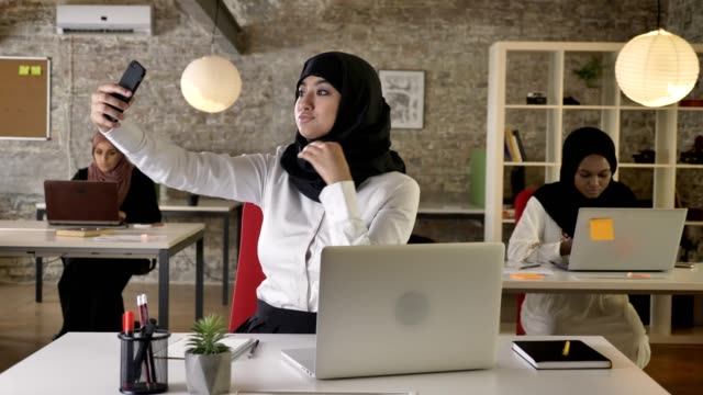 Tres-jóvenes-mujeres-musulmanas-en-hijab-oficina-moderno,-mientras-que-otra-mujer-musulmana-tome-selfie-con-smartphone