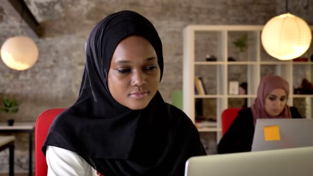 Porträt-des-jungen-schwarzen-muslimischer-Frauen-Hijab-arbeiten-am-Laptop,-konzentriert,-zwei-charmante-Damen-sitzen-im-modernen-Büro