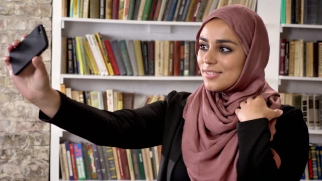 Portrait-of-young-muslim-pretty-women-in-hijab-taking-selfie-then-looking-at-phone-and-watching-photos-in-library,-smiling