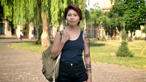 Joven-estudiante-asiática-con-el-tatuaje-del-pie-con-la-mochila-y-mirar-en-la-cámara-en-el-parque-cerca-de-la-Universidad,-seguro