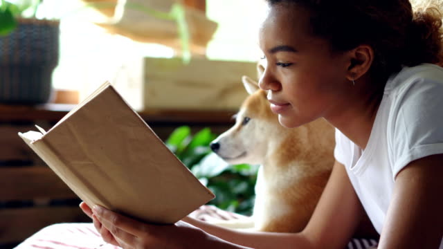 Close-up-shot-of-smart-African-American-woman-reading-book-in-free-time-lying-on-bed-with-her-pedigree-dog-with-large-window-and-green-plants-in-background.