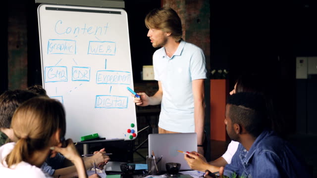 Männlichen-Trainer-arbeitet-mit-Whiteboard-auf-Chart-zeigen-und-sprechen,-während-Mitarbeiter-lernen-und-sitzen-am-Tisch-im-Büro-sprechen.