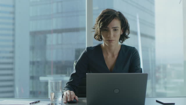 Schönen-erfolgreichen-weiblichen-CEO-arbeiten-auf-einem-Laptop-in-ihrem-modernen-sonnigen-Büro-mit-Stadtbild-Fenster-anzeigen-Starke-weibliche-Führungskraft.
