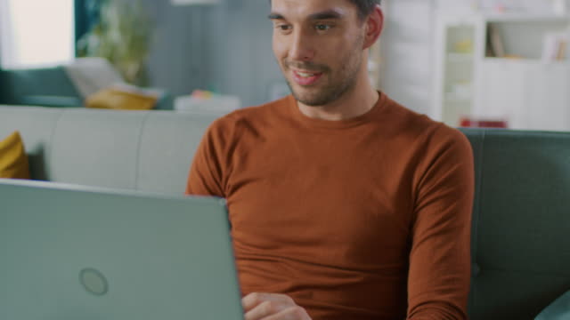 Handsome-Man-Uses-Laptop-Computer-while-Sitting-on-Sofa-at-Home.-Man-Working,-Browsing-Through-Internet-from-His-Cozy-Living-Room.