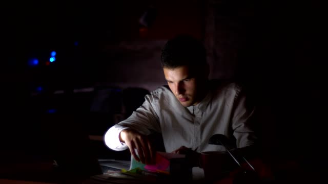 Shooting-of-dark-office,-light-from-screen-ar-shining-on-his-face-while-he-is-sitting-looking-at-computer-and-checking-papers,-holding-them-with-focused-confident-face,-indoor-illustration