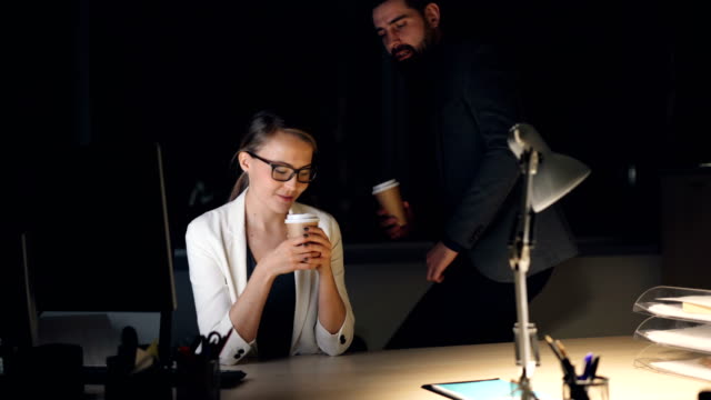 Fürsorgliche-männlicher-Kollege-bärtigen-Mann-bringt-Take-away-Kaffee-zu-müde-junge-Frau,-die-spät-in-der-Nacht-am-Computer-arbeiten.-Mädchen-und-junge-sprechen-lächelnd-und.