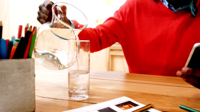 Man-pouring-jug-of-water-in-glass-while-using-mobile-phone