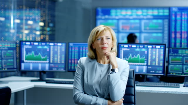 Stock-Market-Leading-Analyst-Thinking-Hard-on-Solving-Financial-Problem.-Behind-Her-People-Working-and-Monitors-Show-Graphs-and-Figures.