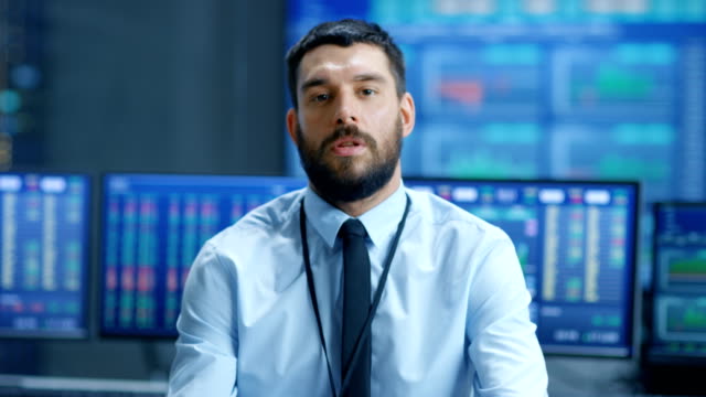 Professional-Stock-Market-Trader-Charismatically-Talks-into-the-Camera.-Behind-Him-Computer-Screens-with-Ticker-Numbers,-Data,-Graphs.