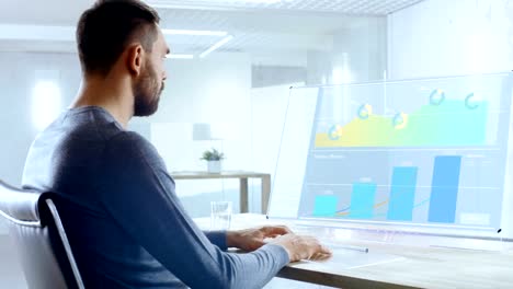 In-the-Near-Future-Office-Worker-Designs-Report-on-His-Transparent-Computer-Display.-Screen-Show-Interactive-User-Interface-with-Statistics,-Graphs-and-Charts.-Office-is-Bright-and-Modern.